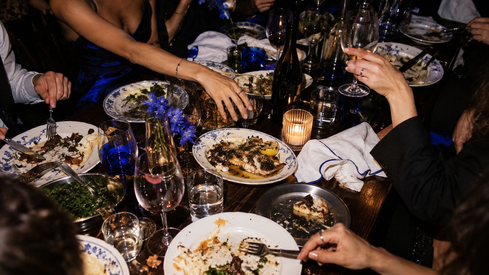 overhead view of a candlelit dinner table at night, hands reaching across shared plates, blue flowers and wine glasses
