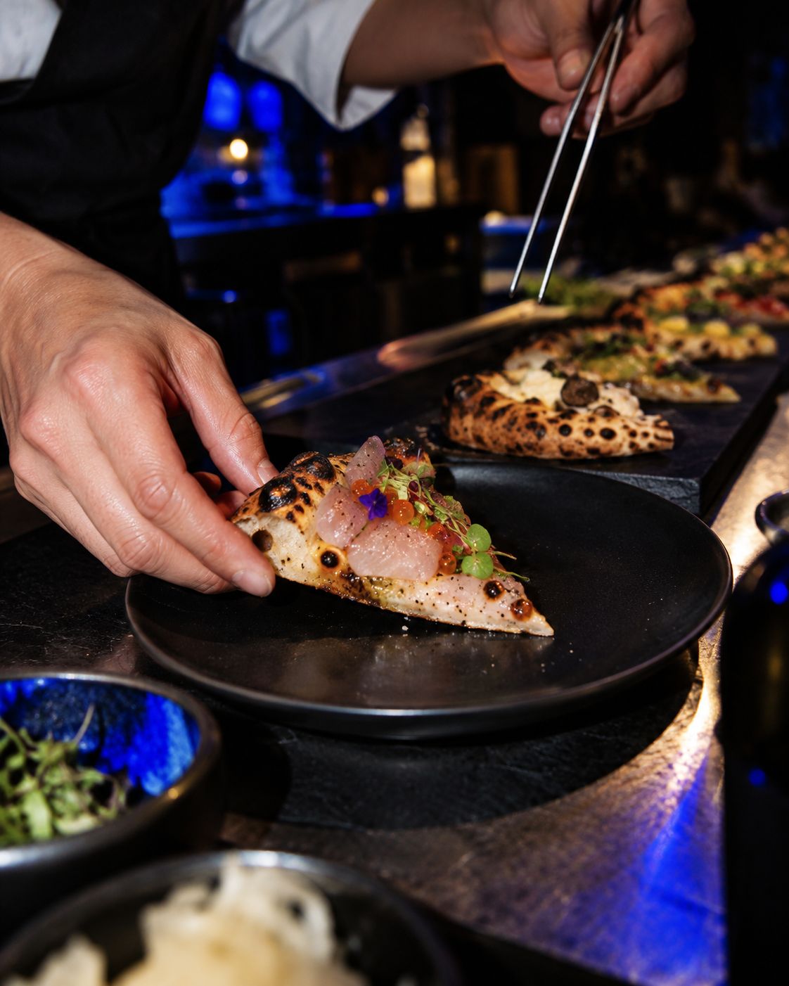 a chef's hands using tweezers to finish a topped slice of pizza on a black plate