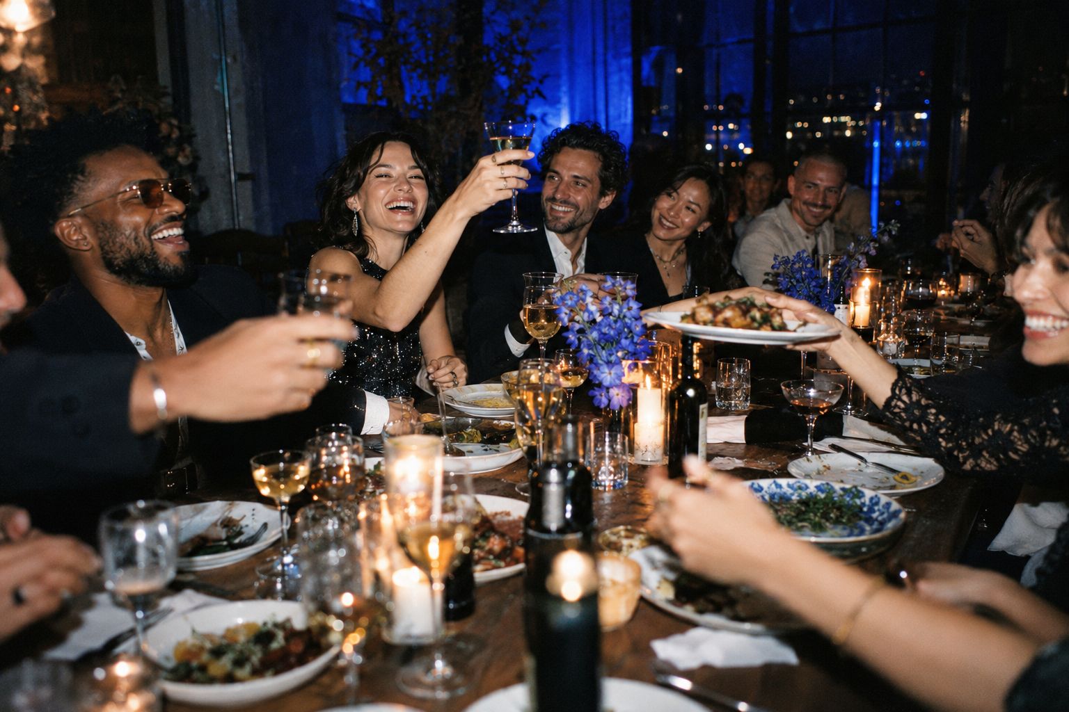 guests laughing and raising their glasses around a long candlelit table under blue light